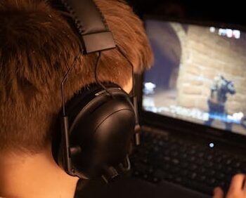 Teenage boy wearing headphones focused on playing a video game on a laptop screen indoors.